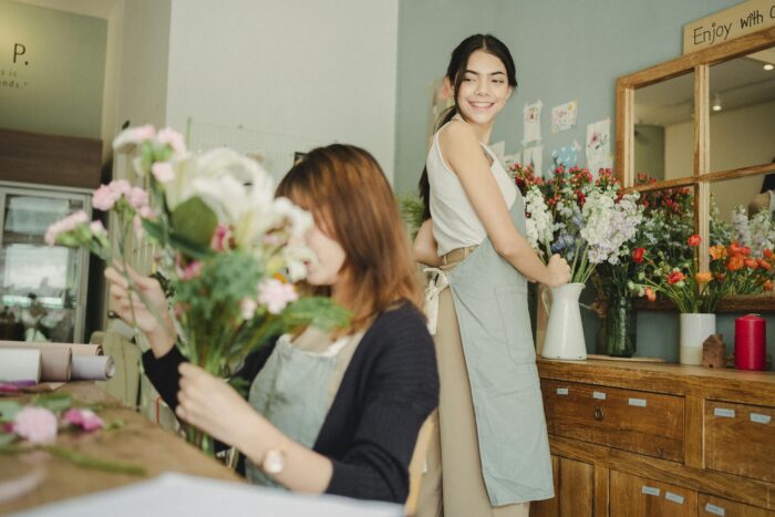 Two female florists creating decorative bouquets in a light-filled workshop. Smiling and in action.